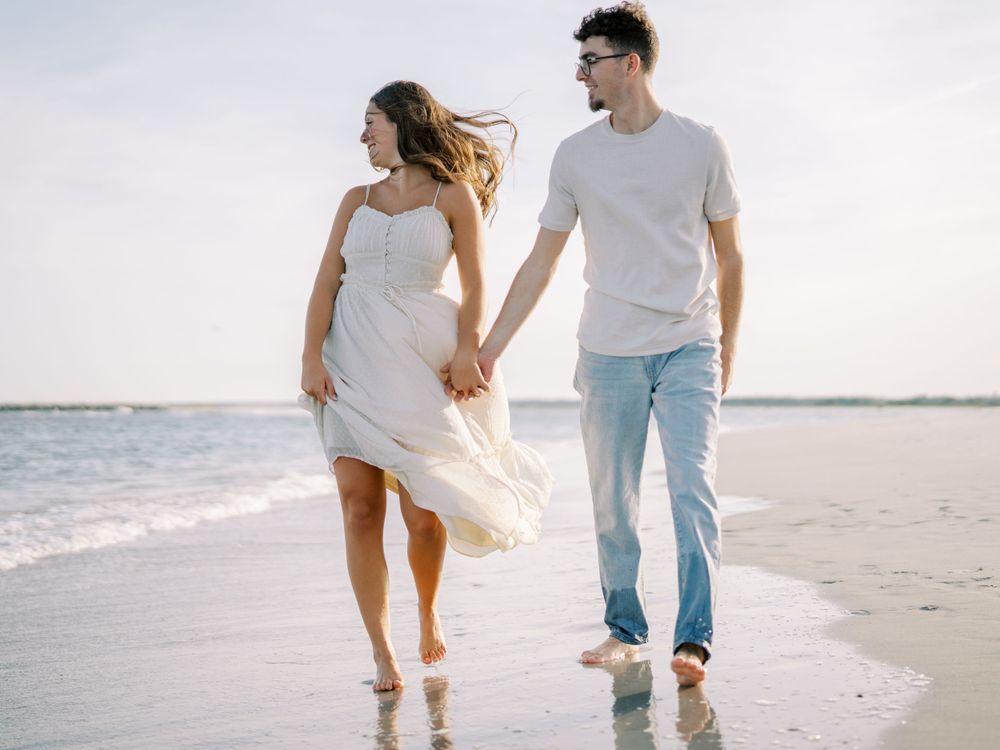 Romantic couple walking hand in hand on a beach with white dress flowing in the breeze.