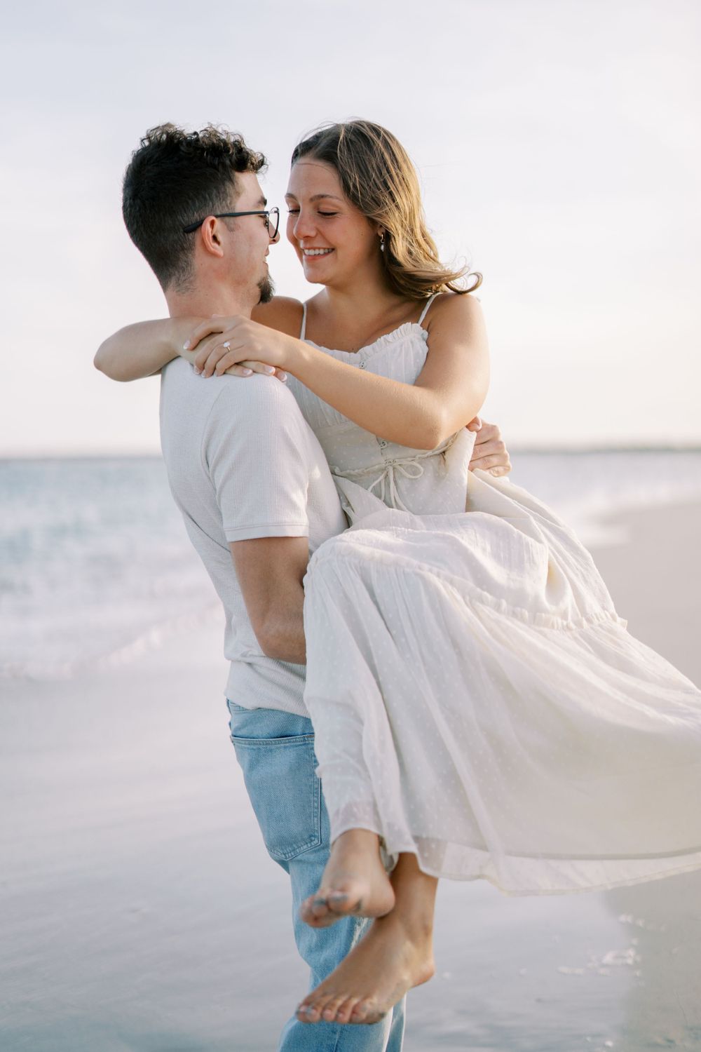 Tender embrace between couple on beach with white flowy dress and light blue jeans.