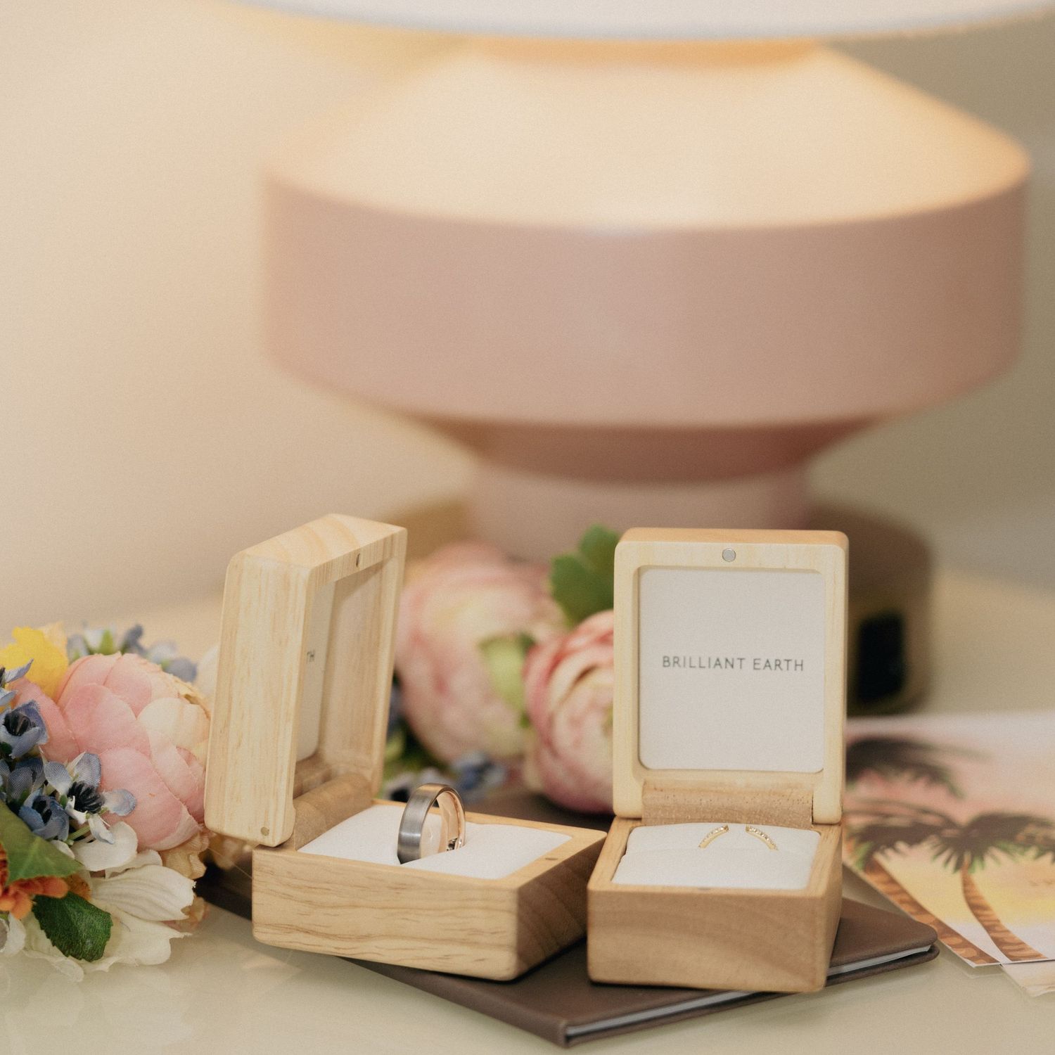Two Brilliant Earth wedding rings displayed in open wooden ring boxes beside a pink lamp and fresh flowers.