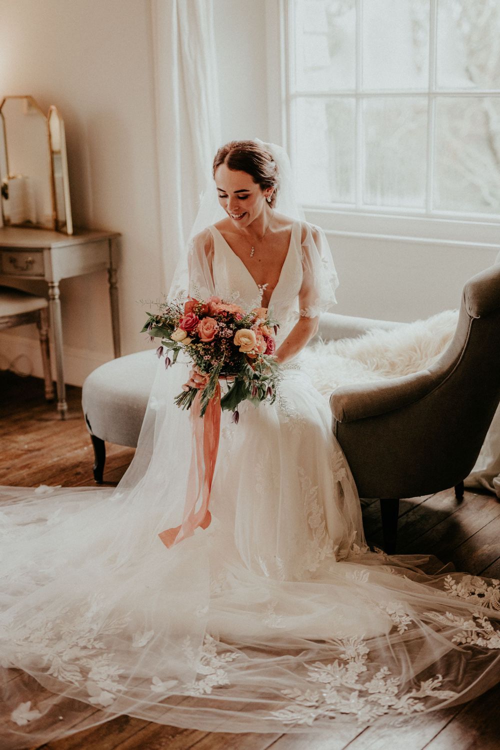 Bride in elegant v-neck gown with flowing tulle skirt sits holding colorful bouquet.