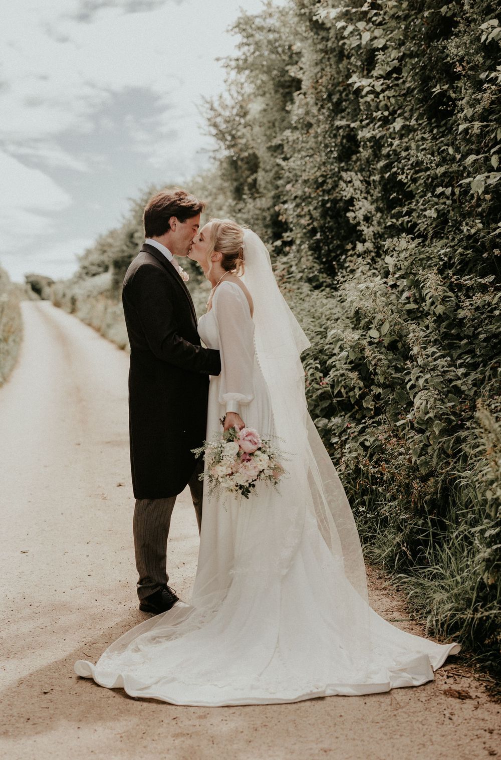 A bride and groom share a kiss on a country lane, with the bride holding a pastel bouquet and wearing a long veil.