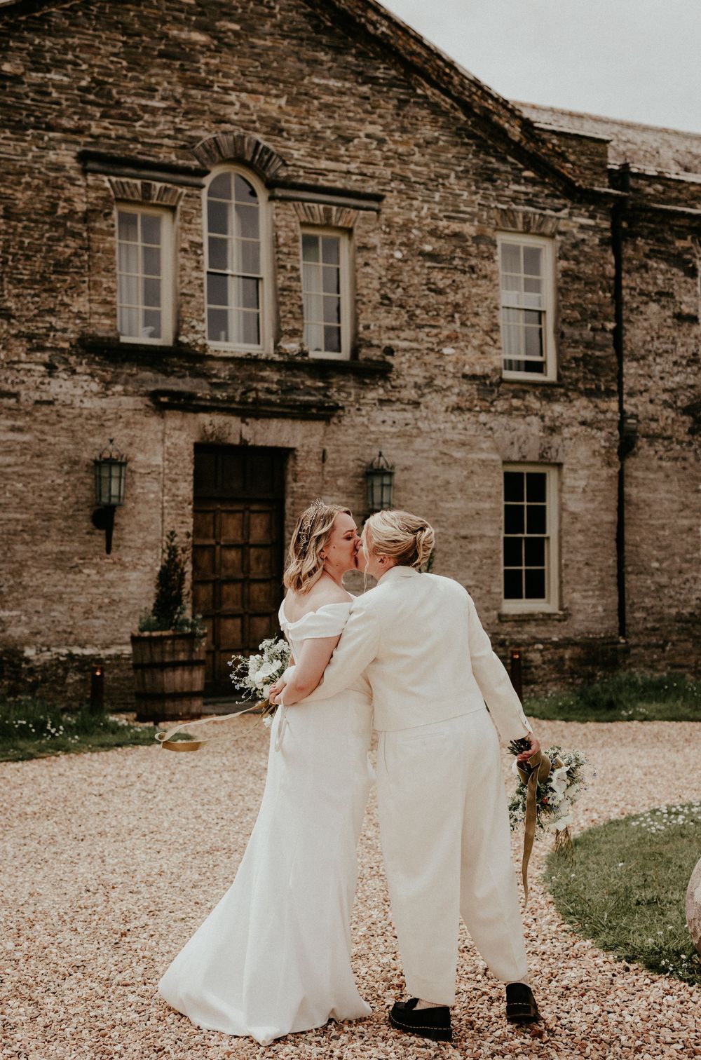 Two brides share a kiss outside a rustic stone wedding venue on a gravel path.