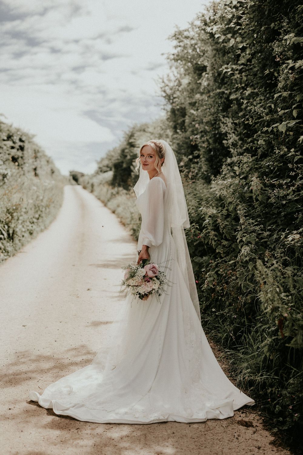 Bride in white gown and veil holds bouquet on a scenic country lane lined with lush green hedgerows.