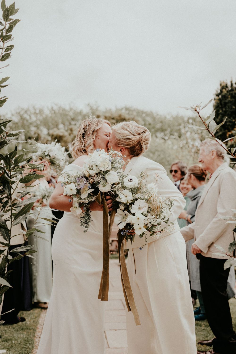 Two brides sharing a kiss at outdoor wedding ceremony, holding white floral bouquets with gold ribbons.