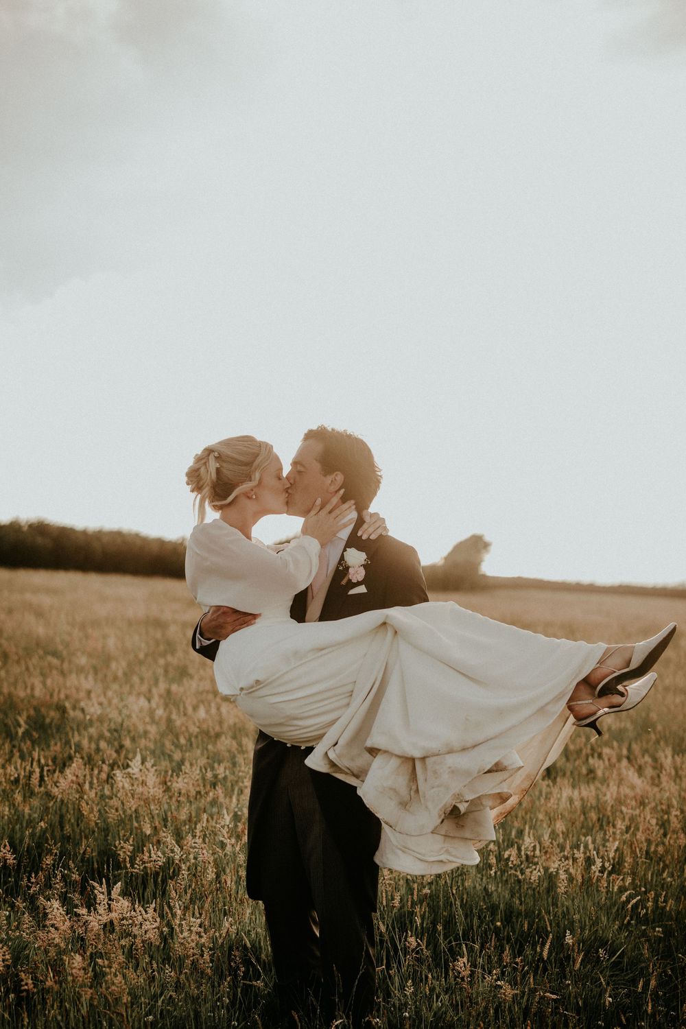 Groom lifts bride in a golden field, sharing a romantic kiss during their outdoor wedding ceremony at sunset.