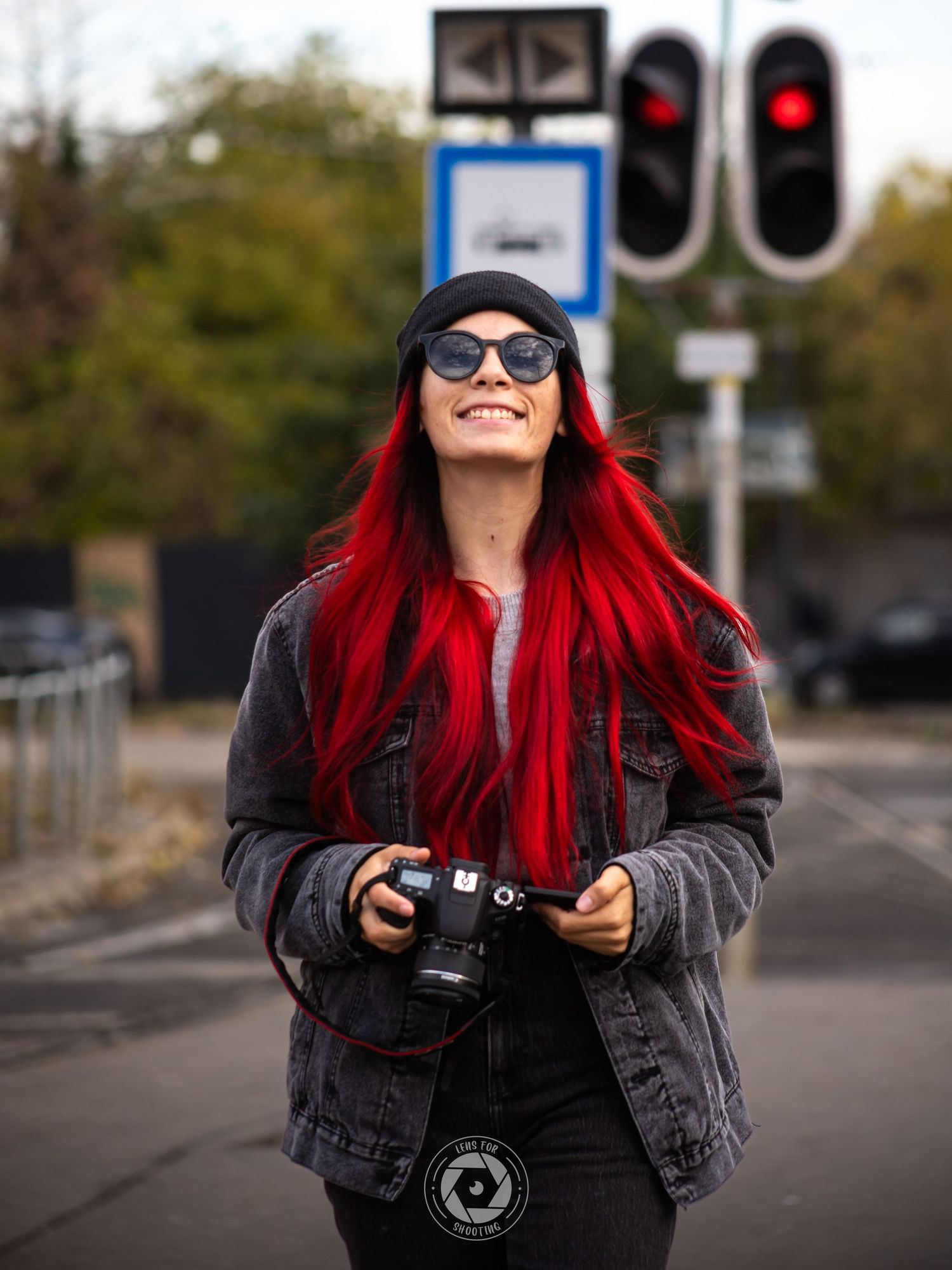 A photographer with vibrant red hair smiles while holding a camera on a city street with traffic lights.