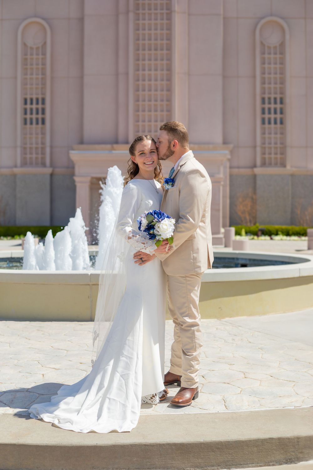 Newlyweds pose by fountain holding blue and white bouquet at ornate building exterior.