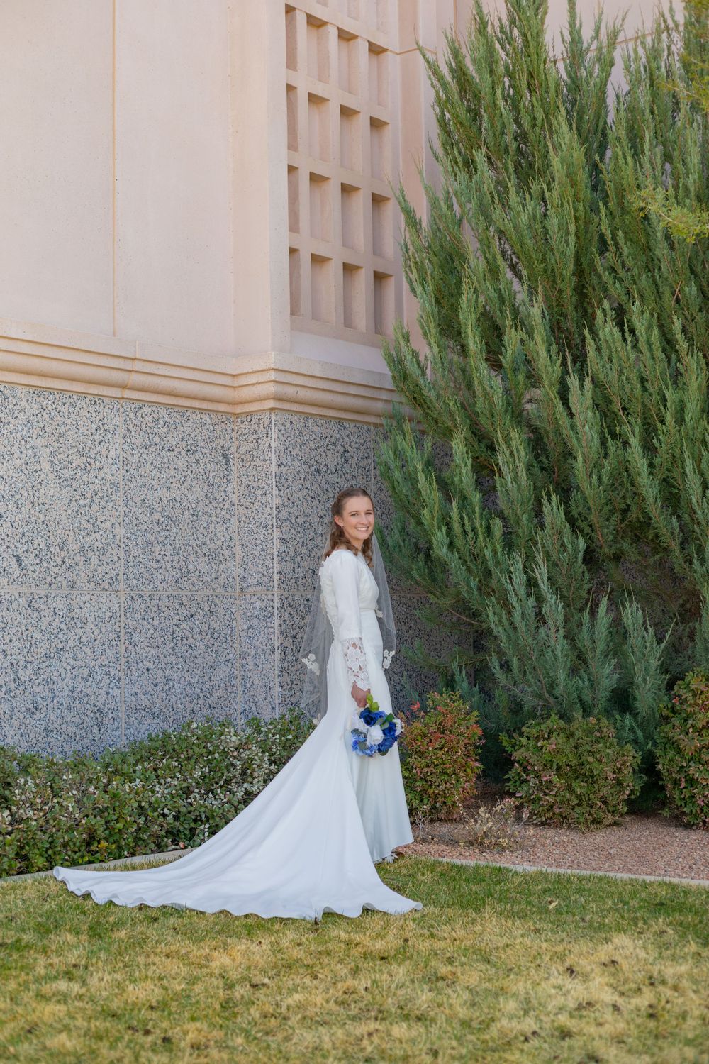 Bride in elegant white dress poses with blue bouquet against architectural backdrop.