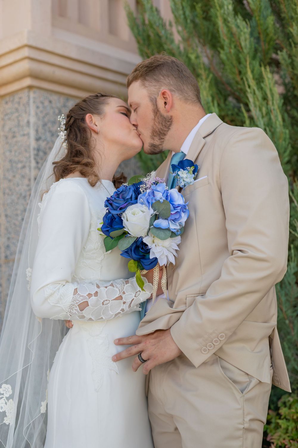 Intimate wedding kiss captured with blue and white bouquet against stone building.