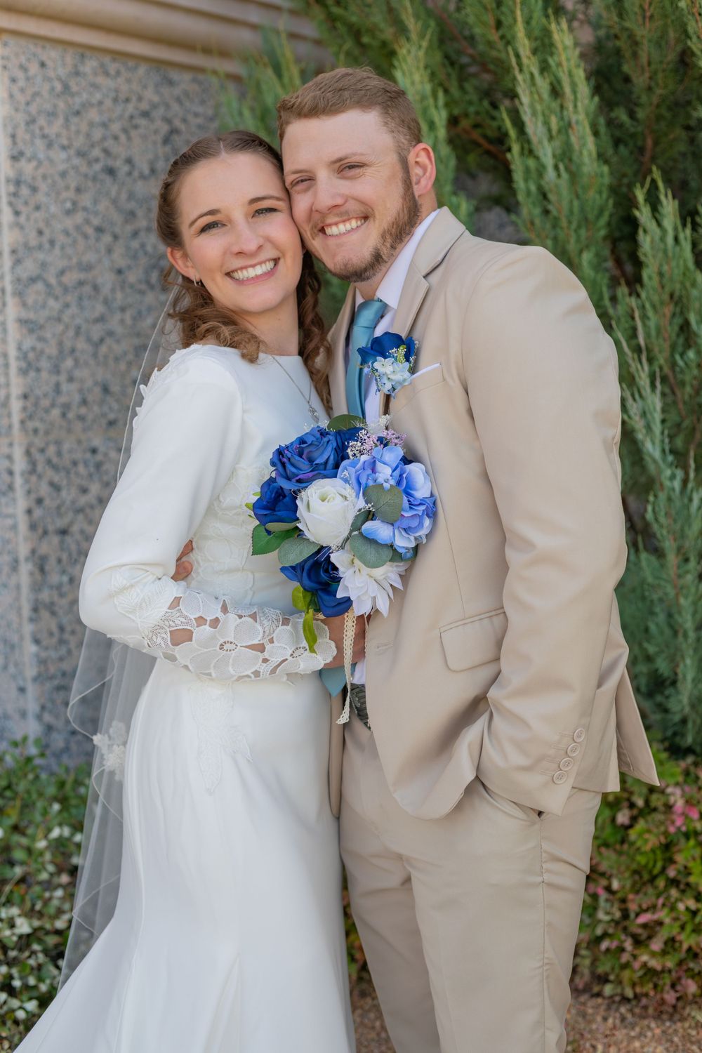 Happy wedding couple poses with vibrant blue floral bouquet outdoors.