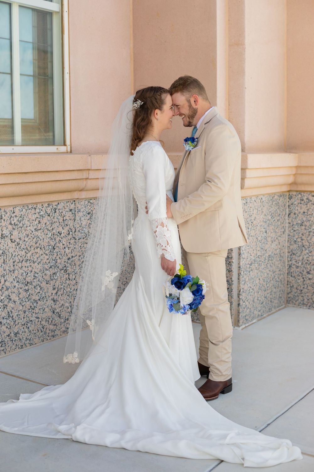 Elegant bride and groom share tender moment against stone building exterior.
