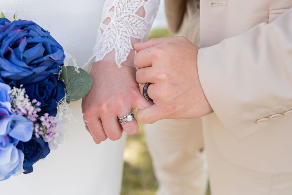 Close-up of wedding rings and lace sleeve details with blue floral bouquet.