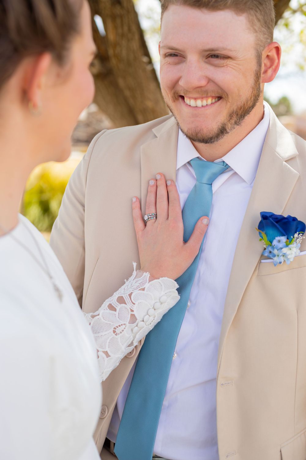 Bride in white lace touches groom's chest as he wears beige suit and blue tie.