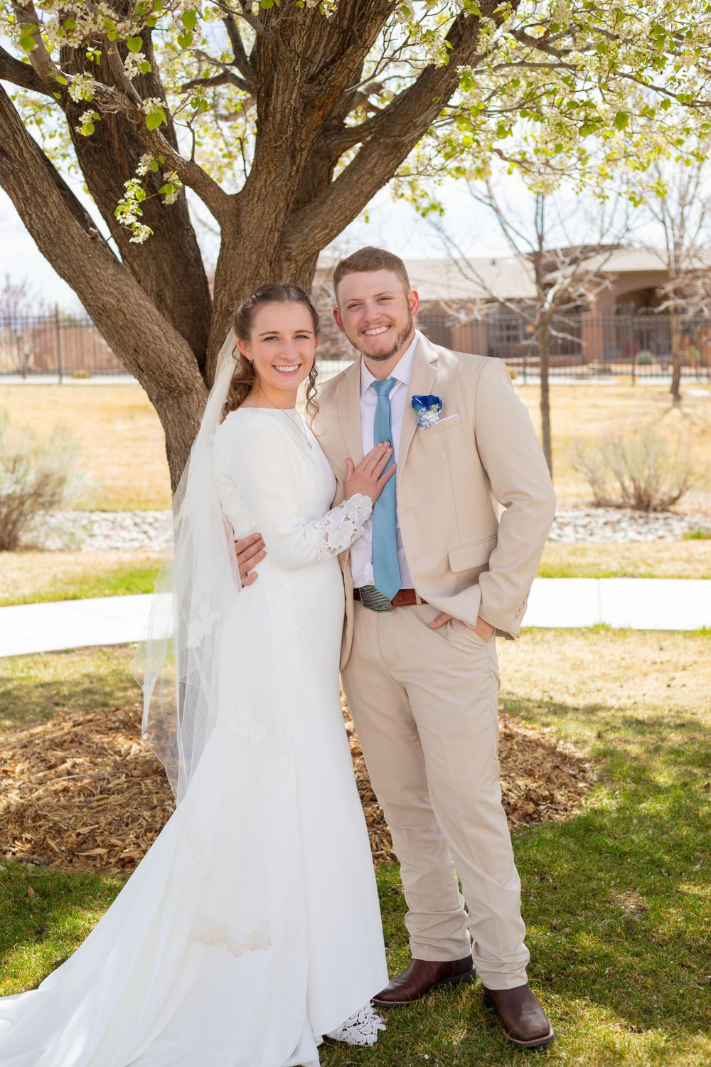Newlyweds pose under large tree during spring wedding in beige and white attire.