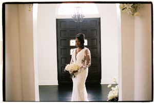 A bride in a fitted white lace gown holds a bouquet while standing in front of a dark door in an elegant hallway.