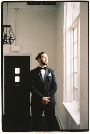 A man in a dark suit stands thoughtfully by a bright window in a minimalist white interior space.