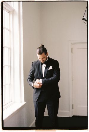 A groom in a black suit checks his phone while standing in a bright room with white walls and windows.
