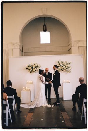 A wedding ceremony takes place in an elegant arched room with white floral arrangements and candles on display.