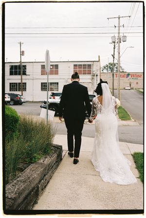 A couple in wedding attire walks away from the camera on a sidewalk with industrial buildings and power lines in background.