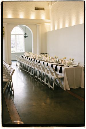 Long banquet table with white linens and chairs set for wedding reception in elegant room with arched window.