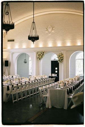 Wide-angle view of a bright wedding venue with arched windows and long banquet tables set for formal dining.