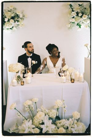 Wedding reception table decorated with white flowers and candles showing a couple sitting at head table during celebration.
