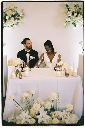 Wedding couple sits at head table decorated with elegant white floral arrangements and candles at reception.