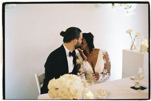 A romantic couple shares an intimate moment at a restaurant table with wine glasses and white decor.