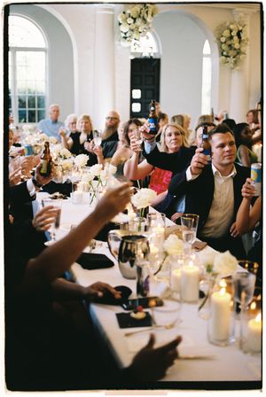 Wedding guests raise their glasses in a toast during an elegant reception with candlelit tables and floral decorations.