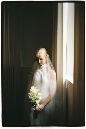 A bride in a white veil holds white lilies against a dark interior with dramatic window lighting.