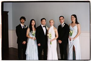 Wedding party members wearing black suits and white dresses pose together in a formal group photo.