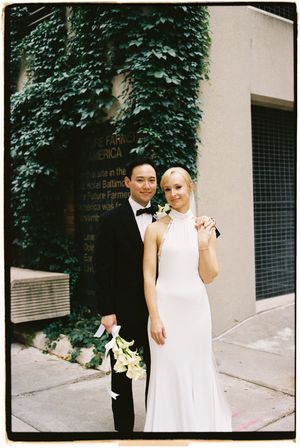 A couple in white attire poses together against an ivy-covered wall on their wedding day.