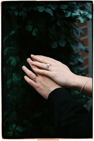 Close-up of hands with wedding rings and bracelet against dark foliage background.