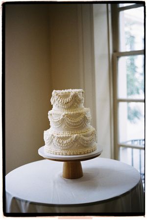 A classic white tiered wedding cake with delicate piped details sits on a white tablecloth near windows.