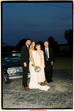 Wedding party gathers at dusk in a gravel parking area near vintage cars.