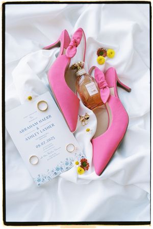 Wedding details flatlay with pink heels, invitation, perfume bottle and rings on white silk fabric.