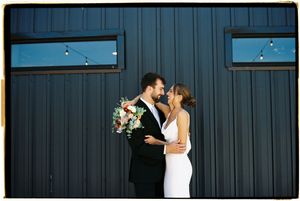 A romantic couple sharing a kiss against a dark blue industrial wall with windows.