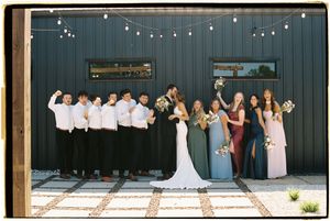 Wedding party poses for photos in front of a dark barn wall with string lights overhead.