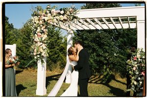 A couple shares a kiss under a white pergola decorated with floral arrangements during an outdoor wedding ceremony.