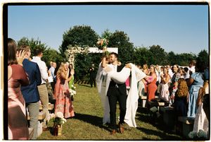 A group of people gathered outdoors for a wedding ceremony on a grassy field under blue skies.