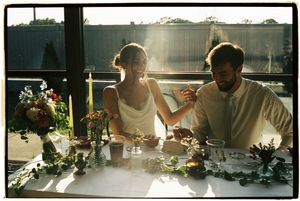 People sit at a long table by a sunlit window, sharing a meal and conversation in a warm, intimate setting.