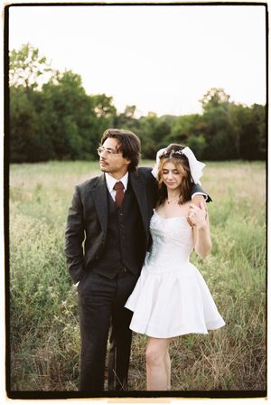 A couple in wedding attire stands together in a grassy field at sunset.