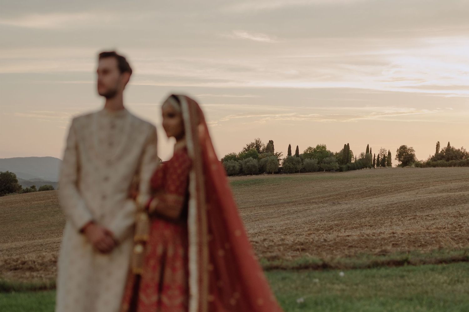 A couple in traditional wedding attire stands in a harvested field at sunset with trees in the background.