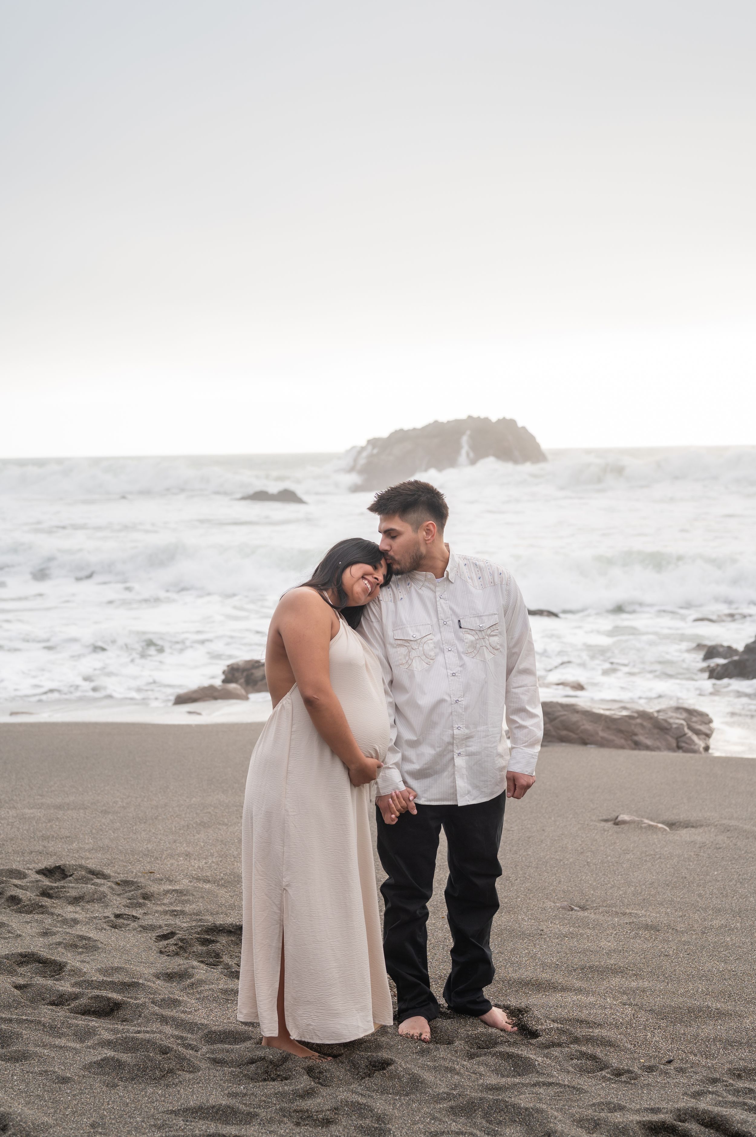 Couple embracing on foggy beach with crashing waves and rocky coastline in background.