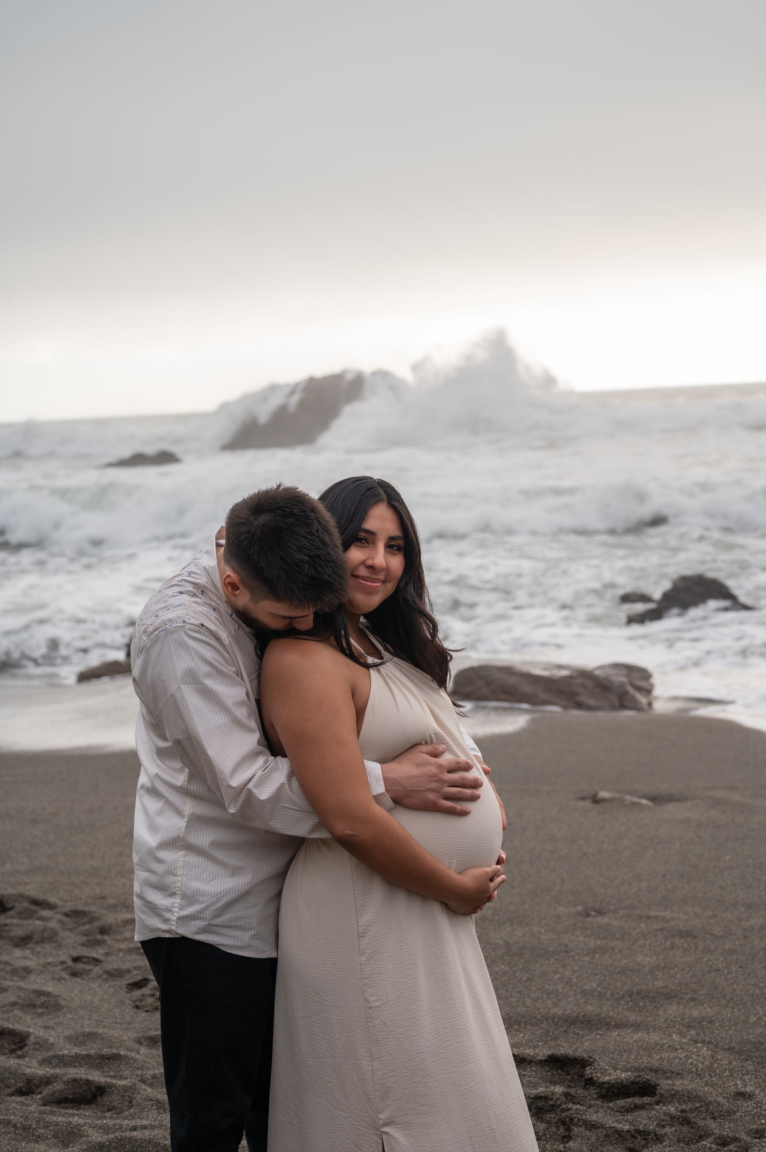 A couple shares an intimate moment on a rocky beach during sunset as waves crash against distant cliffs.
