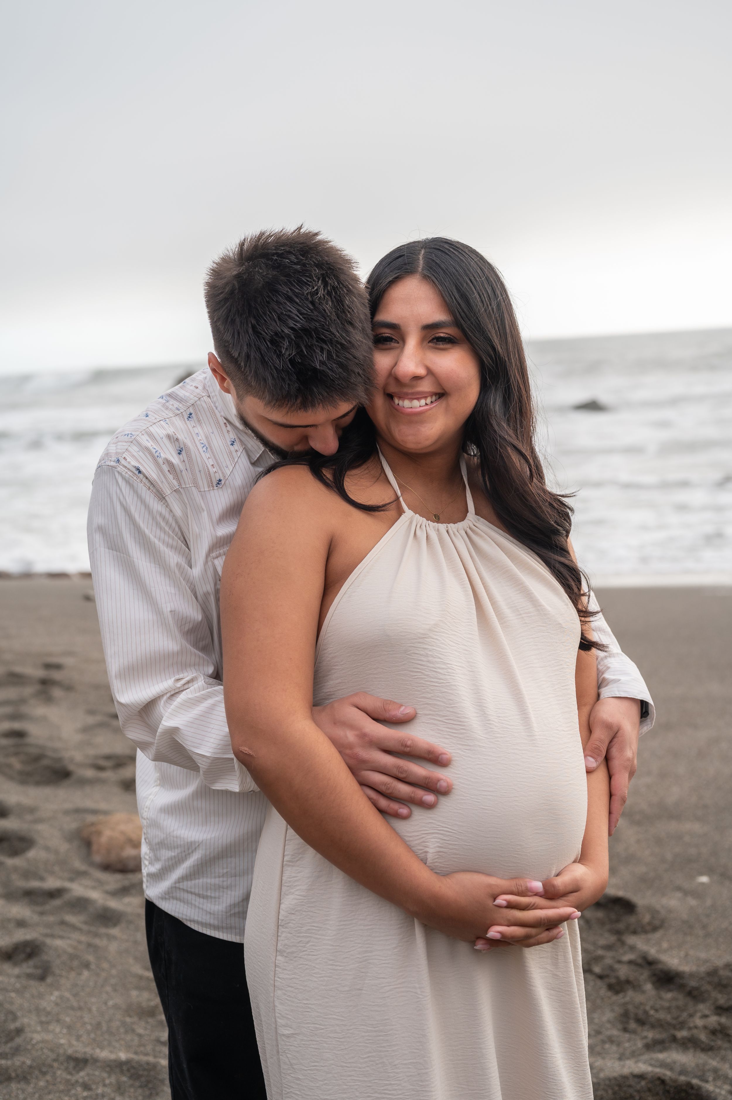 A couple embraces on a cloudy beach while celebrating their pregnancy, with the woman wearing a flowing white dress.