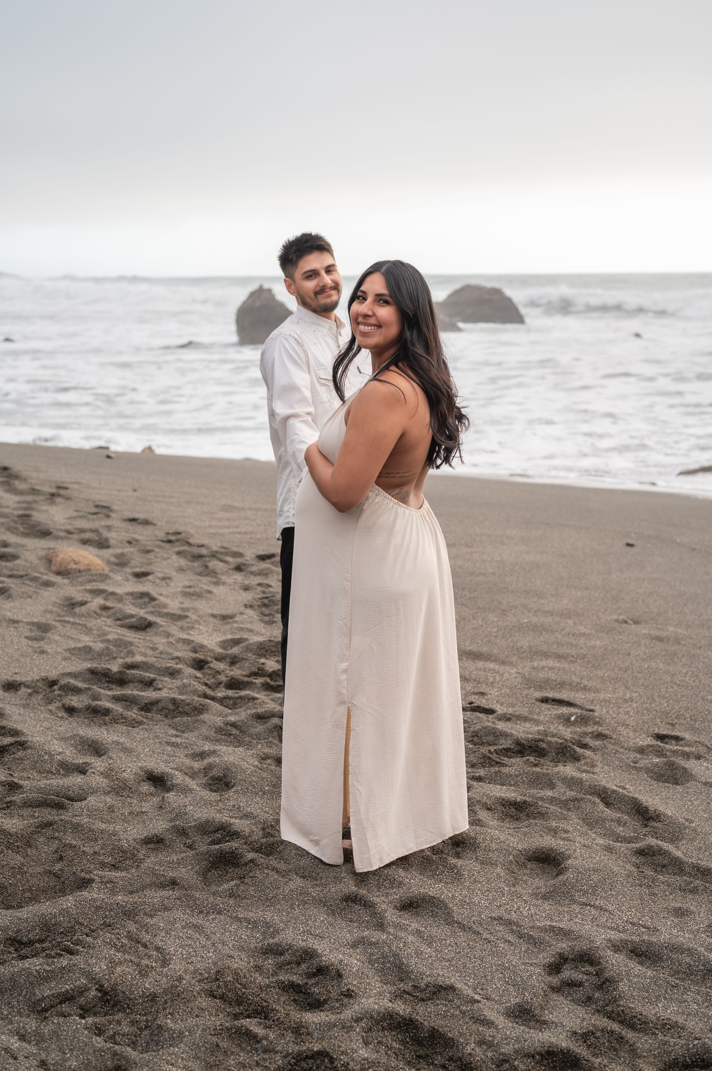 A couple in white attire shares a romantic moment on a misty beach with ocean waves in the background.
