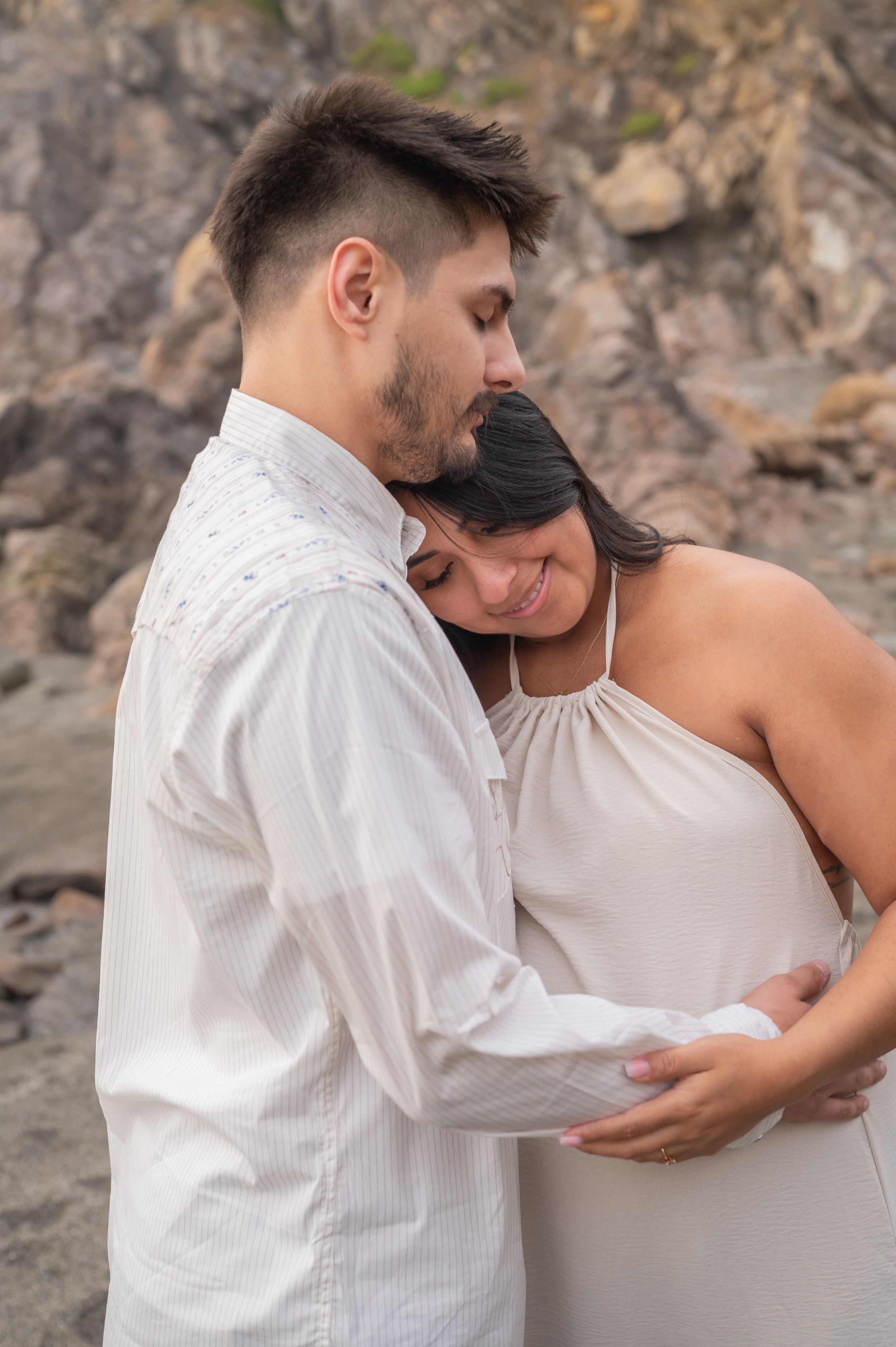 An expectant couple shares an intimate moment on a rocky beach as they tenderly cradle her baby bump.