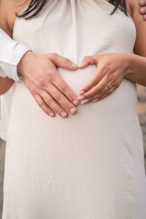Close-up of hands forming a heart shape over a pregnant belly in a white textured dress.