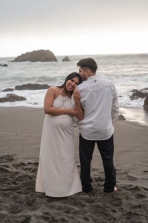 A romantic couple shares a tender moment on a rocky beach at sunset with waves crashing in the background.